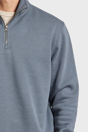 A person is wearing the Academy Brand 1/2 Zip Sweat in light blue with a white undershirt visible at the neckline. The image focuses on the upper torso and part of the neck, emphasizing the sweatshirt's premium brushed fleece material and detailed zipper.