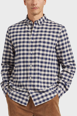 A person dressed in an Academy Brand Aberdeen Shirt, made of blue and white plaid yarn dye mid-weight check fabric, stands with their hands in their pockets. The shirt showcases a classic collar, long sleeves, and heavy enzyme softness. They have paired it with brown pants against a plain white background.