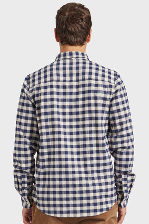 A person with short brown hair is shown from the back, wearing an Academy Brand Aberdeen Shirt. The long-sleeved shirt flaunts a blue and white checkered pattern, made of yarn dye mid-weight check fabric that boasts heavy enzyme softness. The individual appears to be standing against a plain, light-colored background.