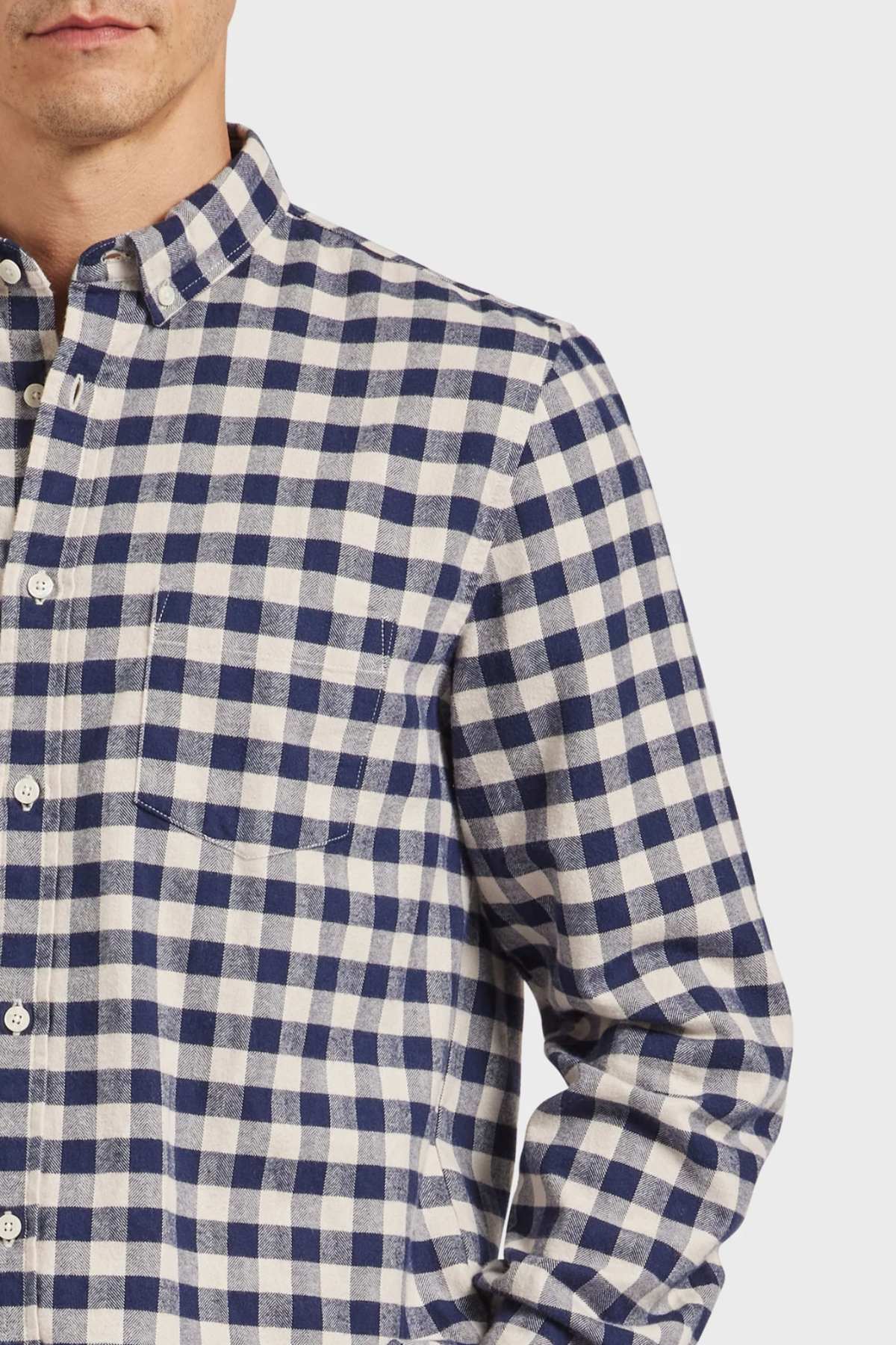 A person is seen wearing the Academy Brand Aberdeen Shirt, a blue and white checked button-up made from mid-weight yarn-dyed fabric. The image focuses on their chest and partially reveals their face, showcasing the shirt's intricate pattern and its heavy enzyme-washed softness.