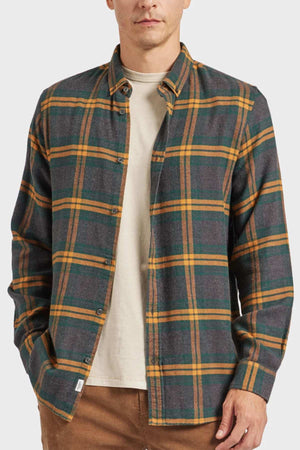 The individual sports an Academy Brand Denver Shirt with green, yellow, and orange plaid stripes over a beige t-shirt, stylishly paired with brown pants against a plain light gray background.