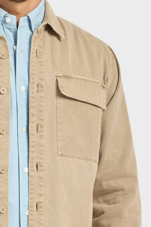 A person wears the Academy Brand Essential Overshirt in tan cotton twill with double chest pockets over a light blue button-up shirt, shown from shoulders to waist against a plain background.