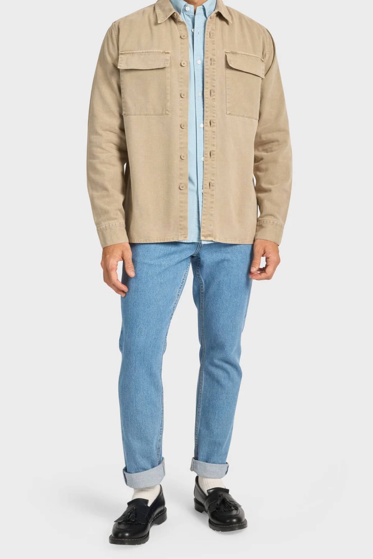 A person is shown from neck to feet wearing the Academy Brand Essential Overshirt in beige cotton twill with double chest pockets over a light blue collared shirt, light blue jeans, white socks, and black loafers.