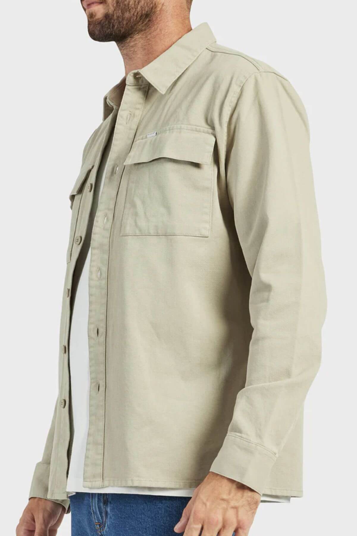 A man is shown from the side wearing an Academy Brand Essential Overshirt in light beige over a white t-shirt, paired with blue jeans. Only the upper part of his body is visible. The durable cotton twill shirt highlights its double chest pockets, with one pocket being buttoned. The background is plain white.