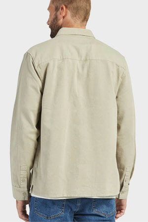 A person with short hair and a beard is standing facing away, wearing the Academy Brand Essential Overshirt in light beige, which features double chest pockets and buttoned cuffs, paired with blue jeans. The background is plain and light-colored.