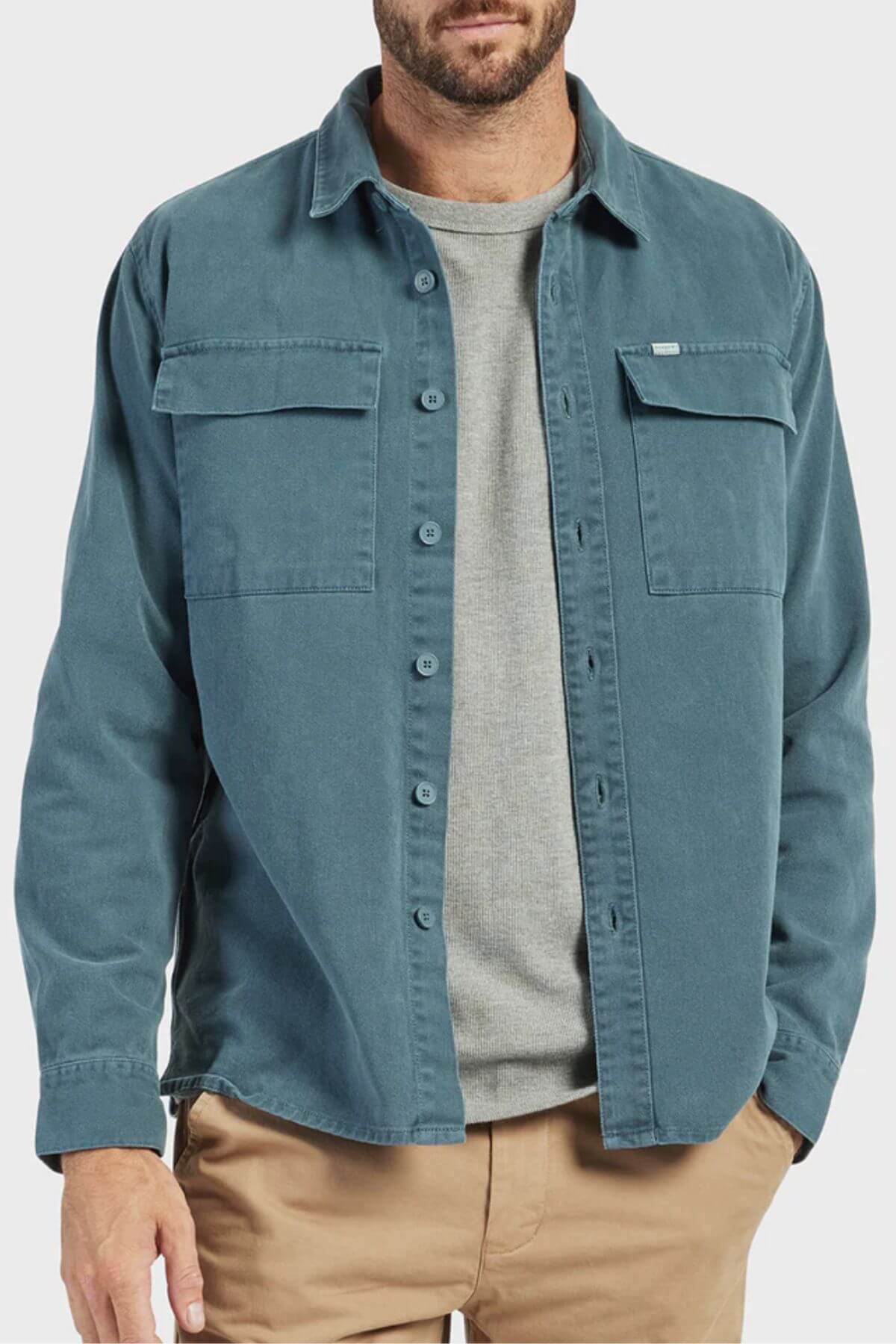 A man wears a teal Academy Brand Essential Overshirt over a gray t-shirt and tan pants, shown from shoulders to thighs against a plain light background.