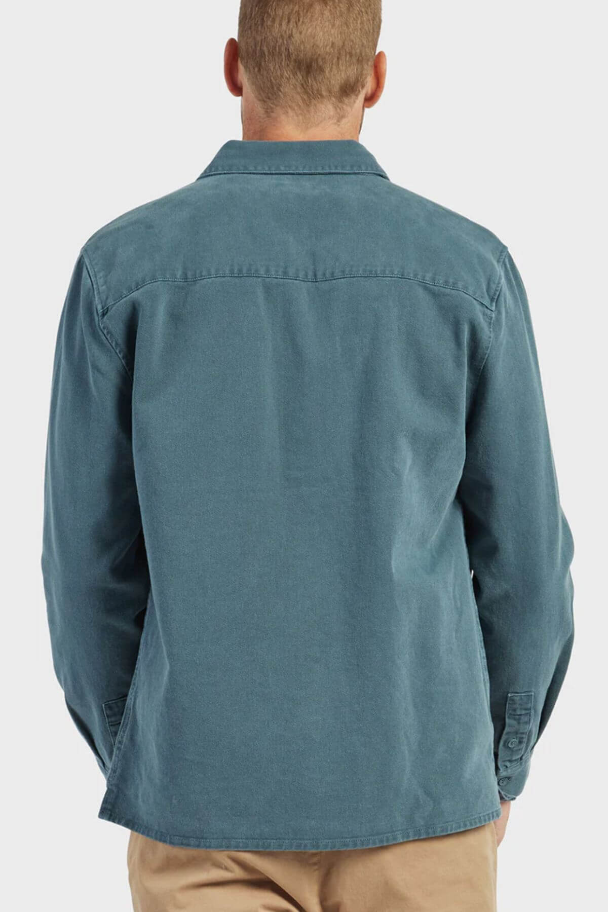 A person with short hair is seen from behind, wearing a teal Academy Brand Essential Overshirt made of cotton twill and beige pants against a plain light background.