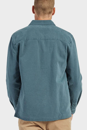 A person with short hair is seen from behind, wearing a teal Academy Brand Essential Overshirt made of cotton twill and beige pants against a plain light background.