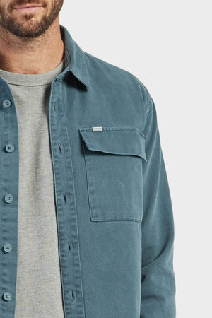 A man wears an Academy Brand Essential Overshirt in teal cotton twill over a light gray ribbed crewneck sweater, shown from shoulders to mid-chest, with part of his beard and mouth visible against a plain background.