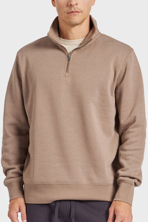 A person in an Academy Brand 1/2 Zip Sweat, taupe over a light shirt and dark pants, featuring ribbed cuffs and hem from premium brushed fleece with a partially open zipper. The background is plain white.
