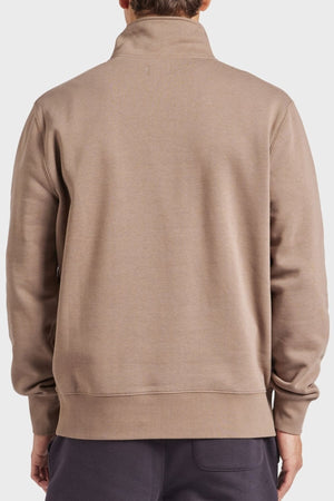 A person is seen from the back wearing an Academy Brand 1/2 Zip Sweat in taupe with a high collar. Made of premium brushed fleece, it features a relaxed fit and is paired with dark pants against a plain white background.