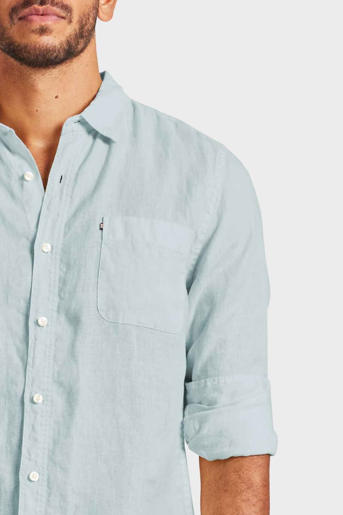 A man wearing the Academy Brand Hampton Linen Shirt in light blue, characterized by rolled-up sleeves and a classic point collar, stands against a plain background. His upper torso is visible along with his face, which features a trimmed beard.