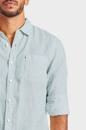 A man wearing the Academy Brand Hampton Linen Shirt in light blue, characterized by rolled-up sleeves and a classic point collar, stands against a plain background. His upper torso is visible along with his face, which features a trimmed beard.