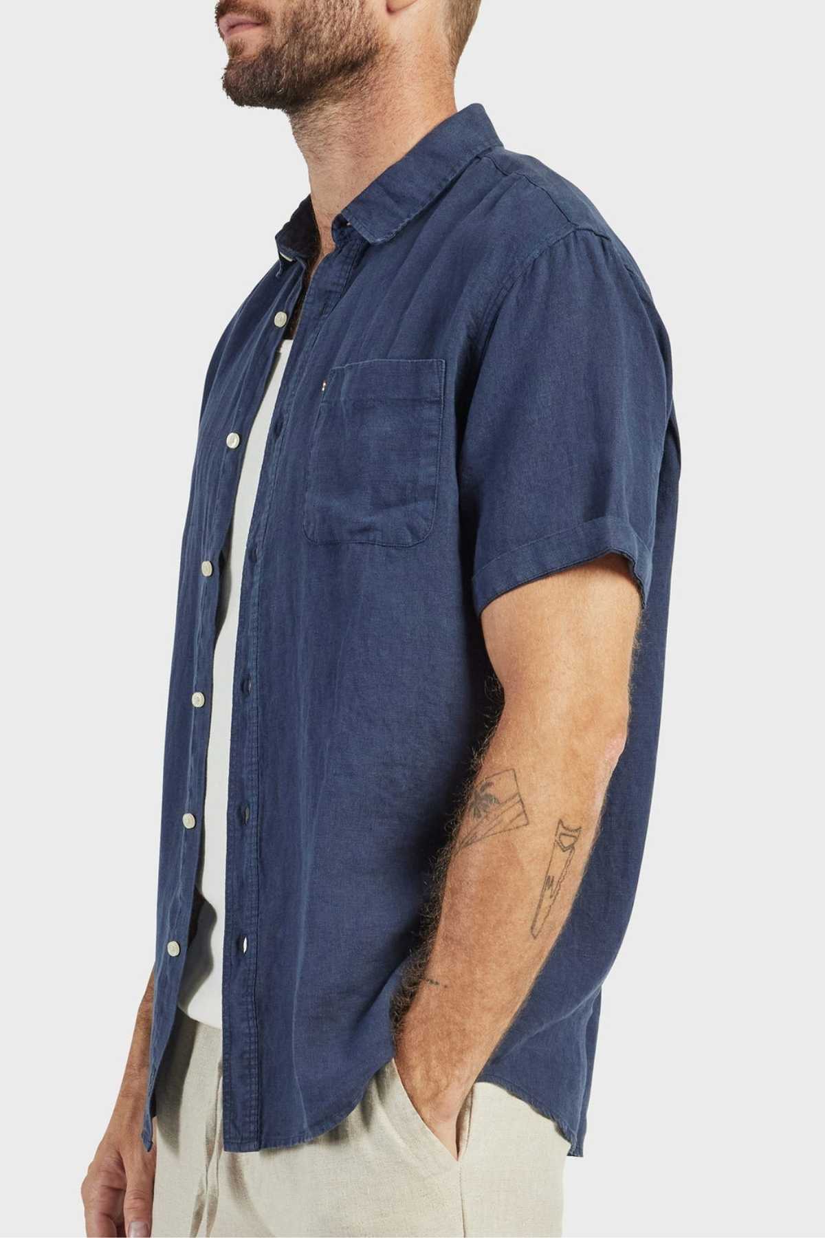 A bearded man with forearm tattoos wears the Academy Brand Hampton Linen Short Sleeve Shirt in navy over a white undershirt and light pants, standing against a plain white background.