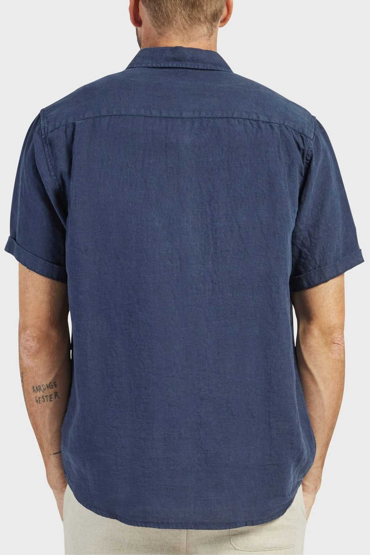 A man with a tattoo on his left forearm, facing away from the camera, wears light pants and an Academy Brand Hampton Linen Short Sleeve Shirt in dark blue. The shirt is 100% linen with a regular fit. The background is plain and light-colored.