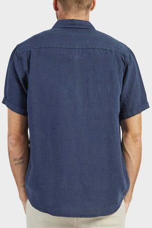 A man with a tattoo on his left forearm, facing away from the camera, wears light pants and an Academy Brand Hampton Linen Short Sleeve Shirt in dark blue. The shirt is 100% linen with a regular fit. The background is plain and light-colored.