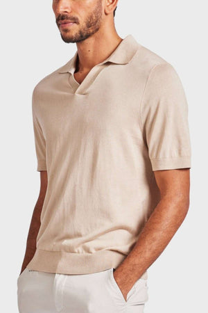 A man is wearing an Academy Brand Irvine Knit Polo in light beige, featuring a vintage-inspired design with a slight V-neck collar. His hands rest in the pockets of his off-white pants. He has a short beard and stands against a plain light gray background, perfectly capturing timeless elegance with this lux blend yarn piece.