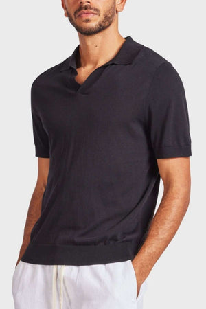A person with short dark hair and a beard, dressed in an Academy Brand Irvine Knit Polo paired with white pants, stands with their hands in their pockets against a plain light gray background.