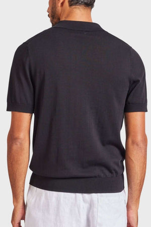 A man with short hair is turned away from the camera, sporting an Academy Brand Irvine Knit Polo—a vintage-inspired black short-sleeved top—and light-colored pants. The backdrop is a plain, light gray tone.