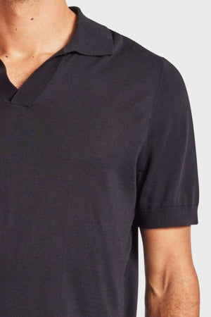 A man is seen wearing a dark Academy Brand Irvine Knit Polo, which features a V-shaped neckline. The photograph captures just the upper torso and part of his left arm, set against a light gray background. This vintage-inspired polo exudes an aura of timeless style and sophistication.
