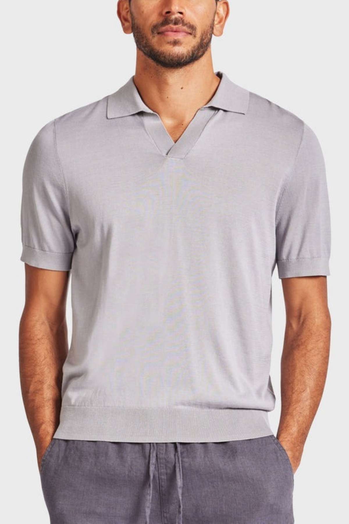 A man stands facing forward, wearing a light gray Academy Brand Irvine Knit Polo with a small V-neck collar and dark gray pants. His hands are tucked into his pockets, and he has a neat beard and mustache. The vintage-inspired polo adds a touch of classic style to his look against the plain, light-colored background.