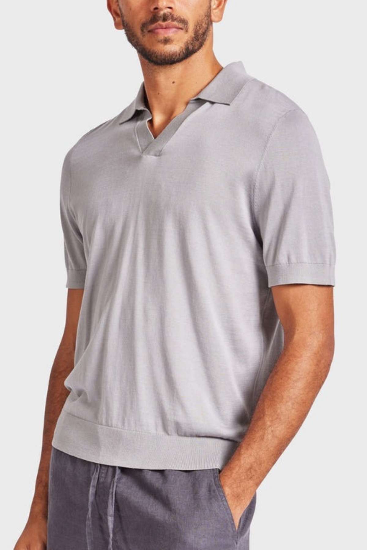 A man is wearing an Academy Brand Irvine Knit Polo in light gray, featuring short sleeves and a collared design made of lux blend yarn. His right hand is in the pocket of his dark gray pants, set against a plain, light-colored background.