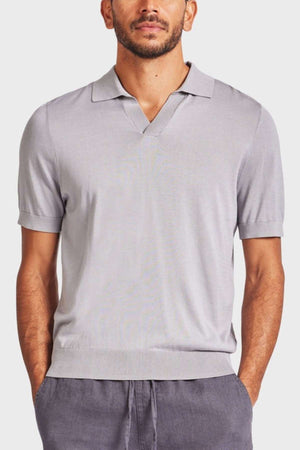 A man stands facing forward, wearing a light gray Academy Brand Irvine Knit Polo with a small V-neck collar and dark gray pants. His hands are tucked into his pockets, and he has a neat beard and mustache. The vintage-inspired polo adds a touch of classic style to his look against the plain, light-colored background.