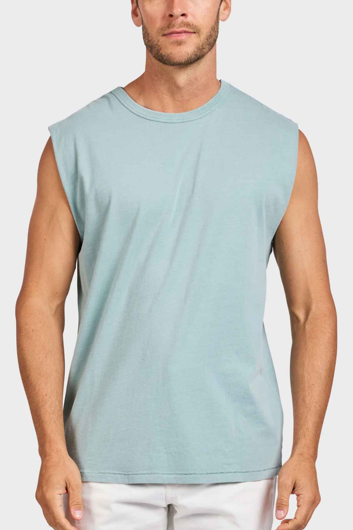 The man is wearing an Academy Brand Jimmy Muscle Tee, a sleeveless light blue heavy-weight sanded jersey, paired with white pants, standing against a plain white background.