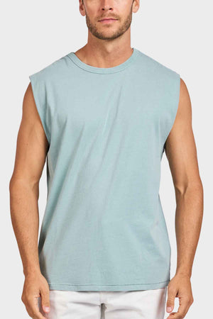 The man is wearing an Academy Brand Jimmy Muscle Tee, a sleeveless light blue heavy-weight sanded jersey, paired with white pants, standing against a plain white background.