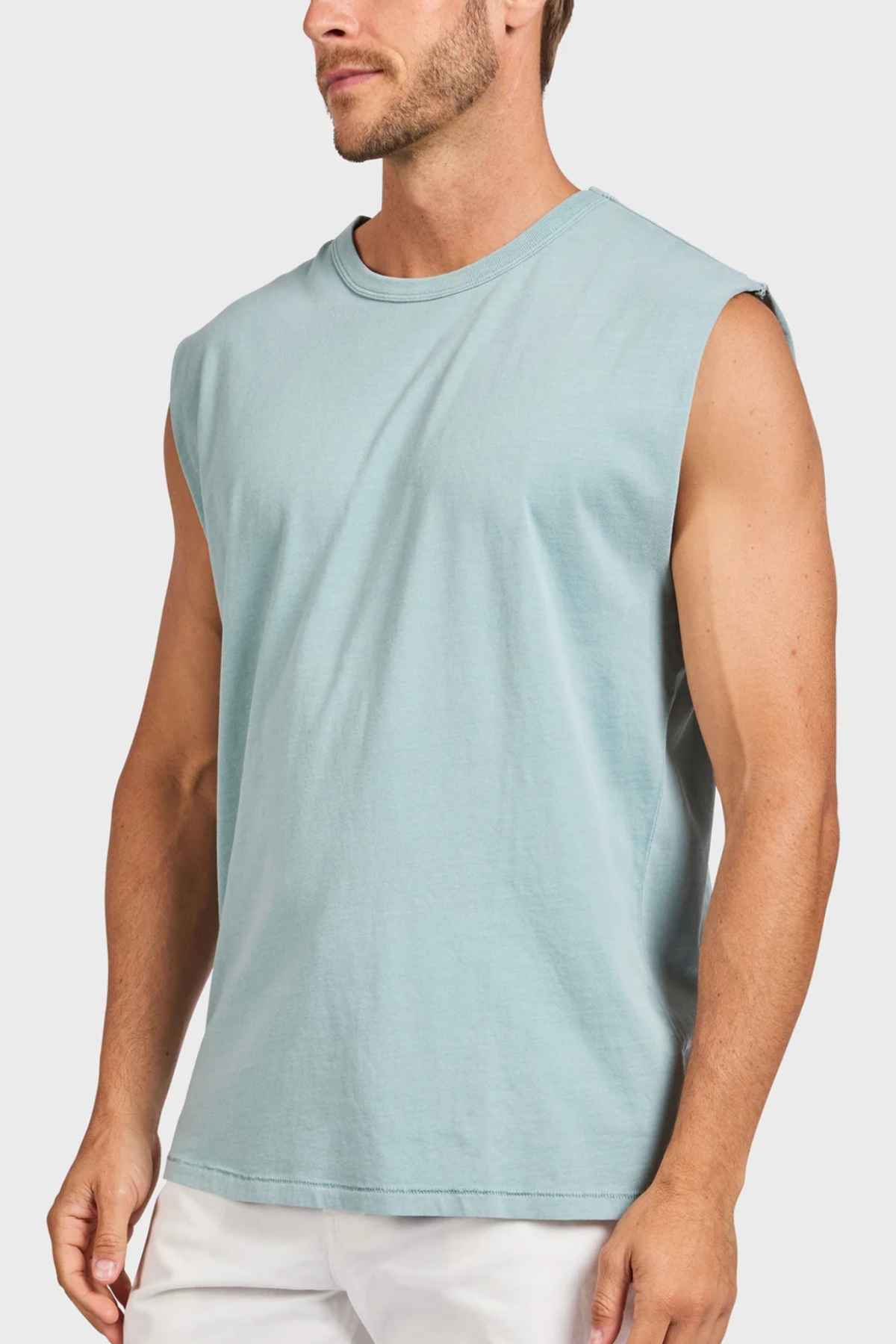 A person wearing a light blue Academy Brand Jimmy Muscle Tee paired with white pants stands against a plain background, exuding a relaxed demeanor with the shirt's comfortable, loose fit made from heavy-weight sanded jersey.