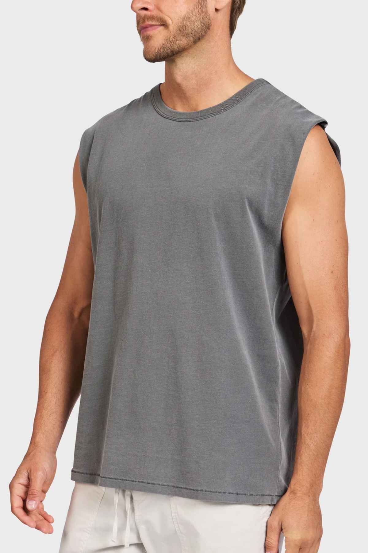 An individual in a sleeveless Academy Brand Jimmy Muscle Tee of gray hue stands against a minimalist background. The T-shirt, made from 100% cotton, is designed to be loose-fitting and casual. The person has short hair and stubble, with their hands resting at their sides.