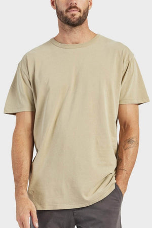 A man wearing an Academy Brand Jimmy Tee in 100% cotton and dark pants, with one hand in his pocket, stands against a light gray background.