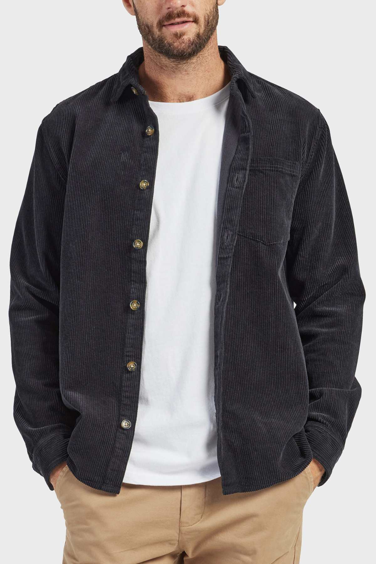 A man dressed in the stylish Academy Brand Lebowski Cord Overshirt, made from mid-weight 100% cotton fabric, layered over a white t-shirt and beige pants. With his hands partially tucked into his pockets and sporting a short beard and mustache, the plain background accentuates his outfit.