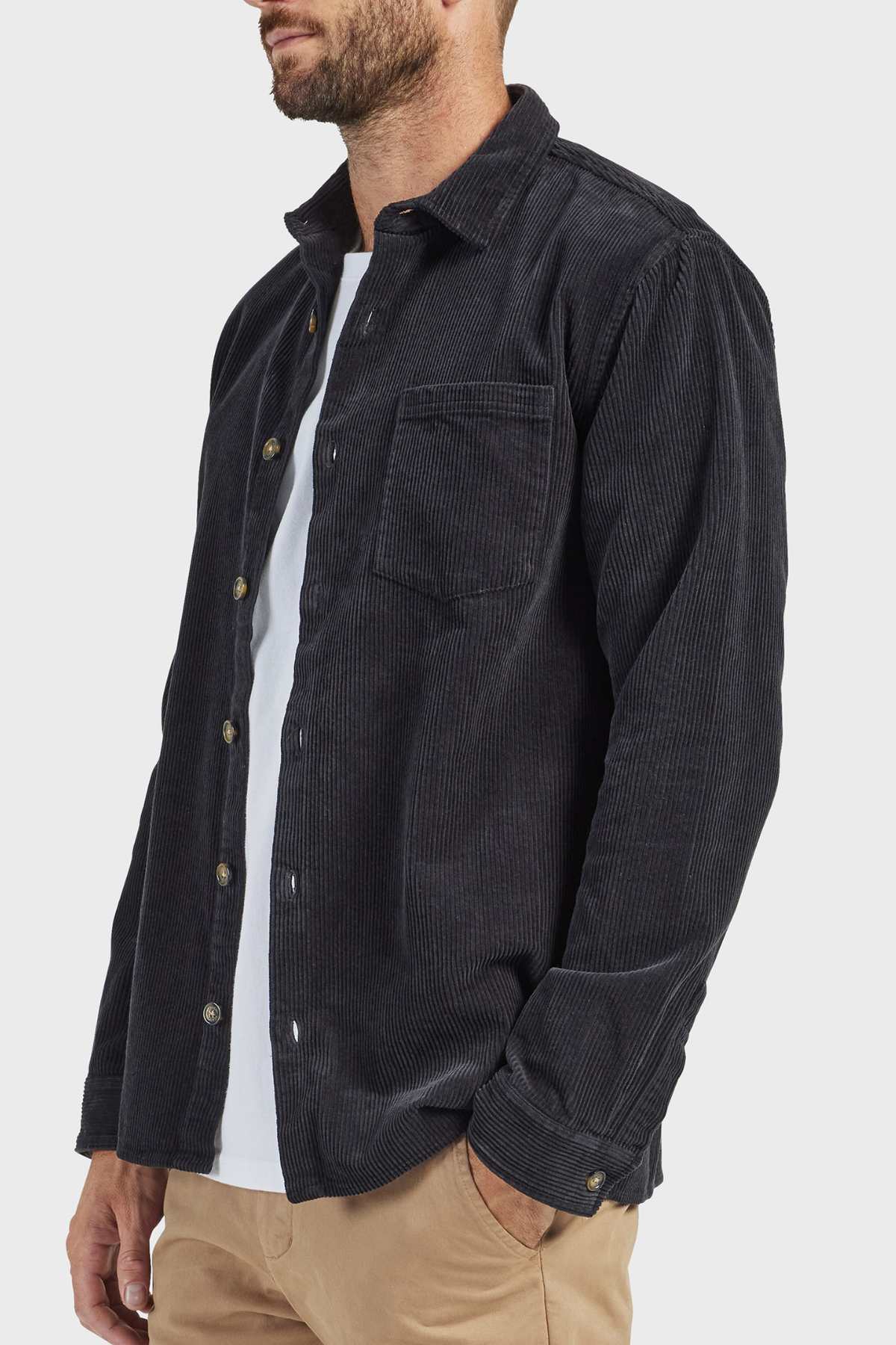 A person is sporting the stylish Academy Brand Lebowski Cord Overshirt in black, crafted entirely from 100% cotton corduroy. The mid-weight shirt, worn unbuttoned over a white T-shirt and paired with beige pants, features a single chest pocket and metal buttons. They are standing with one hand casually placed in their pocket against a plain light-grey background.