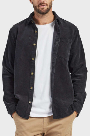 A man dressed in the stylish Academy Brand Lebowski Cord Overshirt, made from mid-weight 100% cotton fabric, layered over a white t-shirt and beige pants. With his hands partially tucked into his pockets and sporting a short beard and mustache, the plain background accentuates his outfit.
