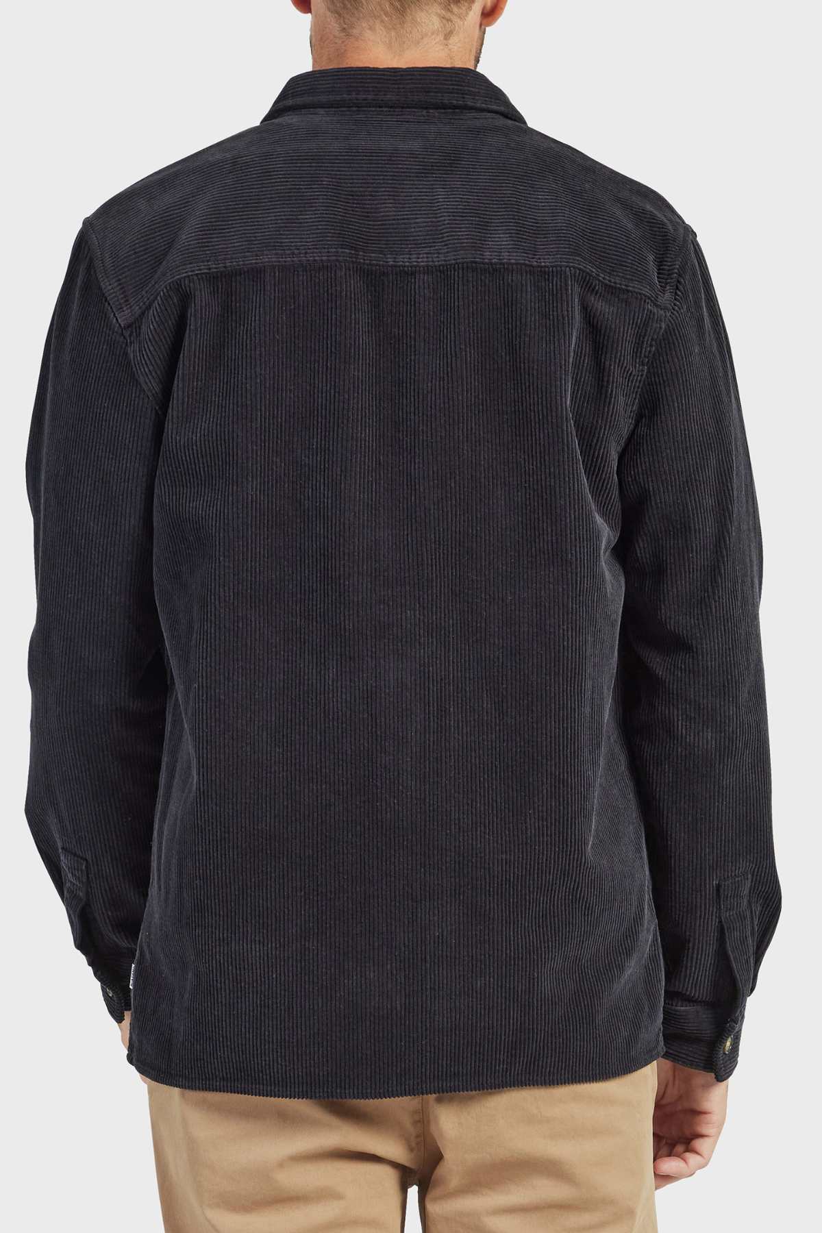 A person is seen from behind wearing the Academy Brand Lebowski Cord Overshirt in stylish black corduroy, crafted from mid-weight fabric with a collar and long sleeves. Their hands are not visible, and they are also wearing light brown pants. The background is plain white.