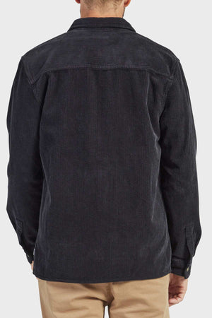 A person is seen from behind wearing the Academy Brand Lebowski Cord Overshirt in stylish black corduroy, crafted from mid-weight fabric with a collar and long sleeves. Their hands are not visible, and they are also wearing light brown pants. The background is plain white.