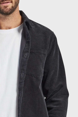 Close-up of a person wearing a partially buttoned Academy Brand Lebowski Cord Overshirt made from mid-weight corduroy fabric over a white T-shirt. The image captures the upper part of the torso and part of the face, highlighting the texture and style of this 100% cotton clothing item.