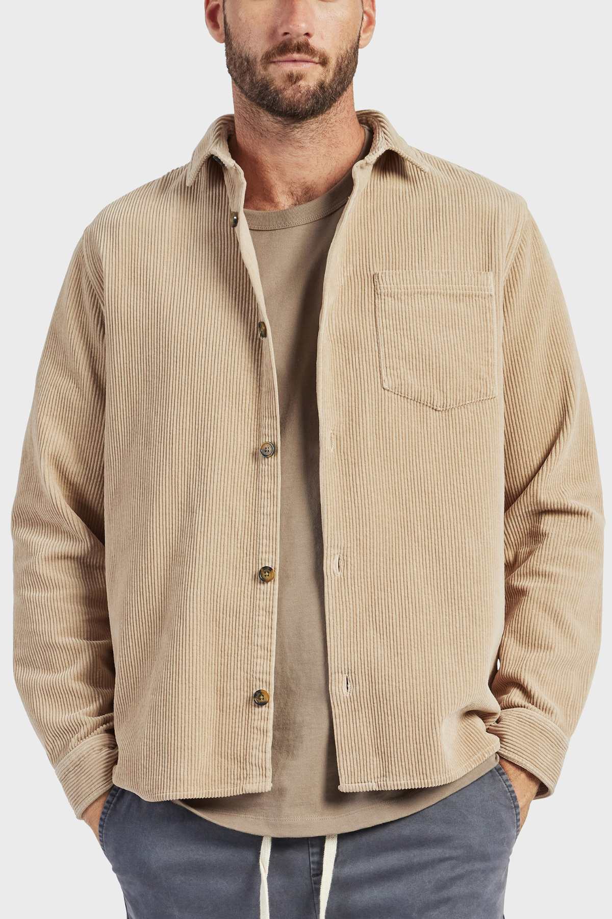 A bearded man stands with his hands in his pockets, wearing an Academy Brand Lebowski Cord Overshirt made of 100% cotton over a taupe t-shirt. He is also wearing dark gray pants with visible drawstrings. The background is plain white.
