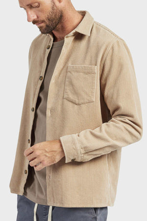 A bearded man wearing the stylish Academy Brand Lebowski Cord Overshirt, made of mid-weight 100% cotton corduroy, over a gray t-shirt looks down while adjusting his sleeve. The plain white background highlights his attire.