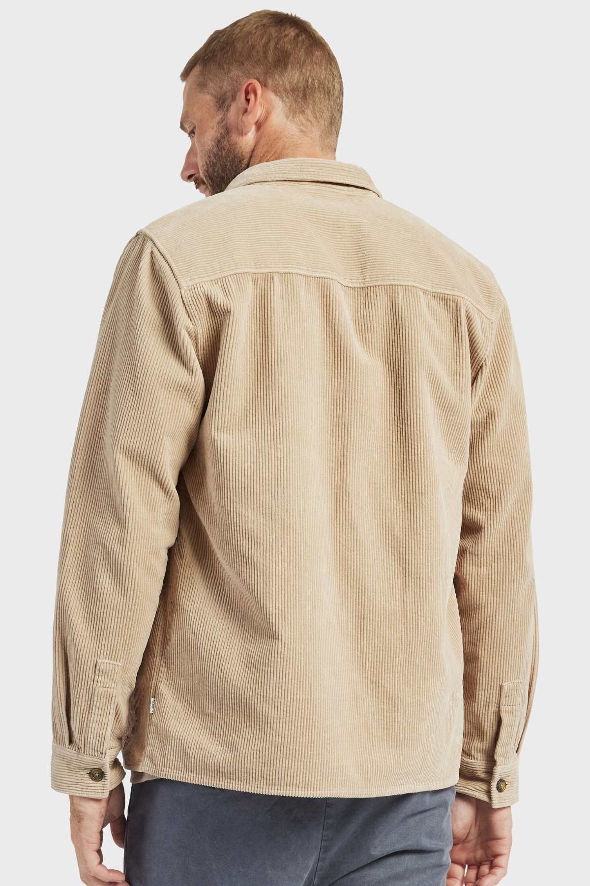 A man with short hair and a beard is seen from the back, wearing an Academy Brand Lebowski Cord Overshirt in a stylish beige color. The long-sleeved shirt, made of mid-weight, 100% cotton, features a textured, vertical ribbed pattern. He pairs it with dark pants against a plain white background.