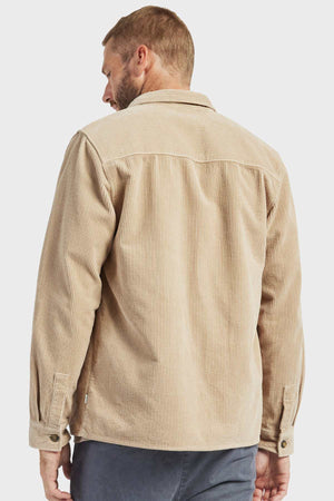 A man with short hair and a beard is seen from the back, wearing an Academy Brand Lebowski Cord Overshirt in a stylish beige color. The long-sleeved shirt, made of mid-weight, 100% cotton, features a textured, vertical ribbed pattern. He pairs it with dark pants against a plain white background.