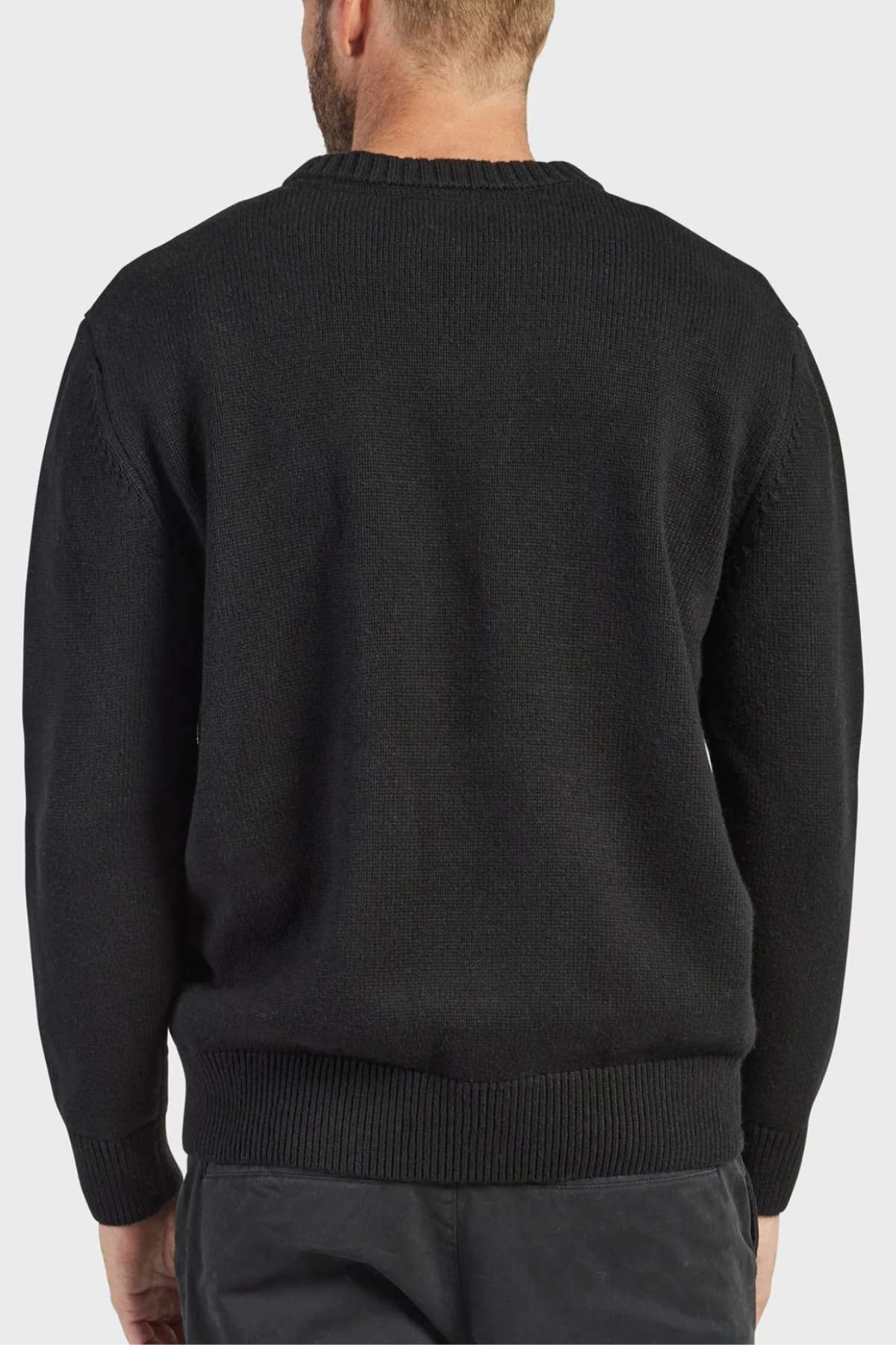 A person with short hair is facing away from the camera, donning a long-sleeved black Academy Brand Malibu Crew sweater paired with dark pants. The image captures the back view of the individual against a plain, light gray background, emphasizing their cozy yet stylish appearance.