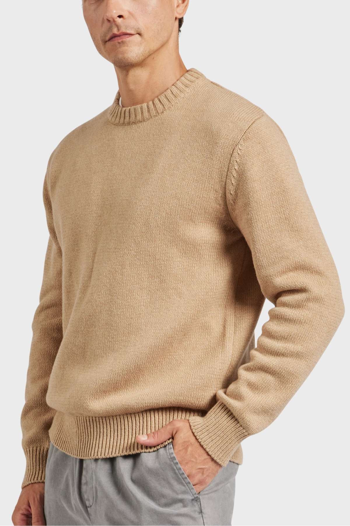 A person wearing the Academy Brand Malibu Crew, a slightly oversized beige long-sleeve sweater with a ribbed crew neck and hemline. The individual has one hand in the pocket of their light gray pants, against a plain white background that emphasizes the high-quality craftsmanship of the sweater.