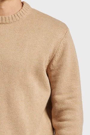 A close-up of a person wearing an Academy Brand Malibu Crew light beige knitted sweater, highlighting the slightly oversized fit. The focus is on the torso and part of the left arm. The sweater features a ribbed neckline and appears cozy and soft against a plain white background.