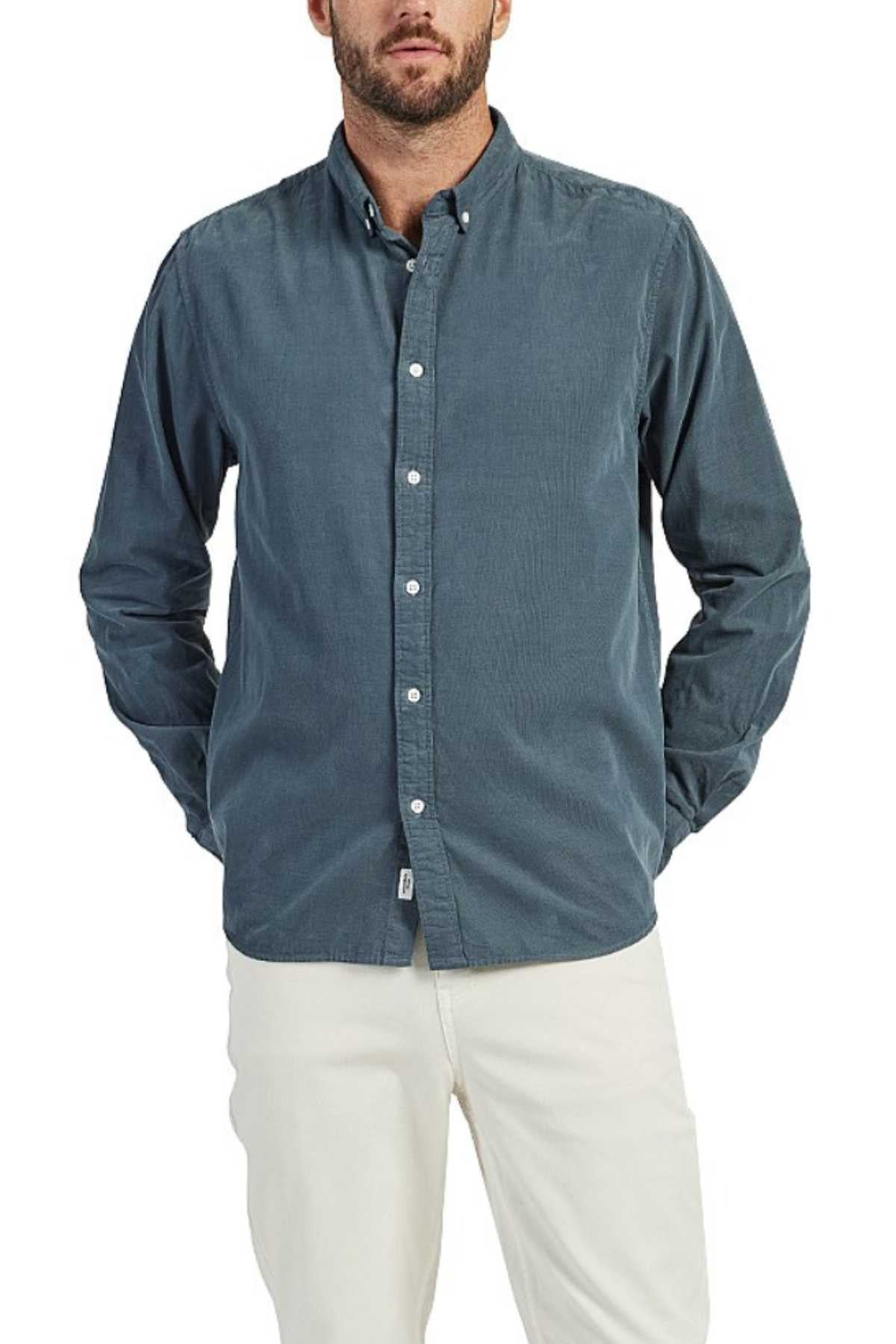 A man is wearing a long-sleeved Academy Brand Micro Cord Shirt in a muted blue color with imitation mother-of-pearl buttons, paired with white pants. His hands are partially in his pockets, and he has a short beard. The background is plain white.