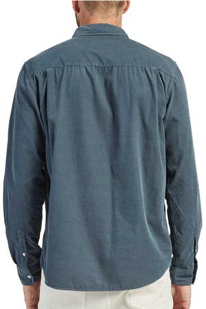 A man is wearing an Academy Brand Micro Cord Shirt in teal, made of 100% cotton. The image captures the back view, revealing its plain and slightly wrinkled design. He pairs it with light-colored pants. The shirt showcases buttoned cuffs, imitation mother of pearl buttons, and a straight back yoke with gathering.