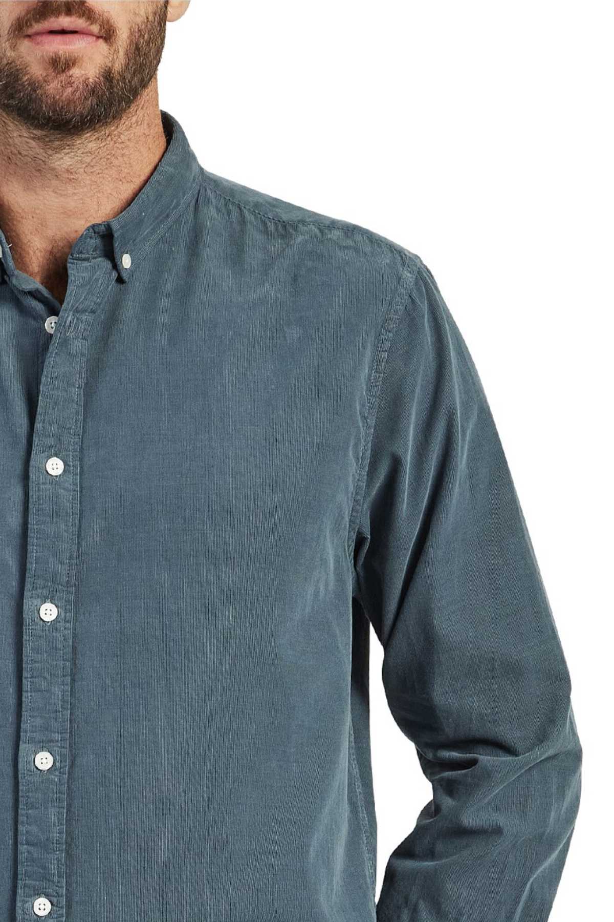 A person wearing the Academy Brand Micro Cord Shirt, a long-sleeved, button-up shirt in teal corduroy with imitation mother of pearl buttons and a collar. Made from 100% cotton, the photo captures the upper torso with the face partially out of frame. The person's right hand is tucked in their pocket.