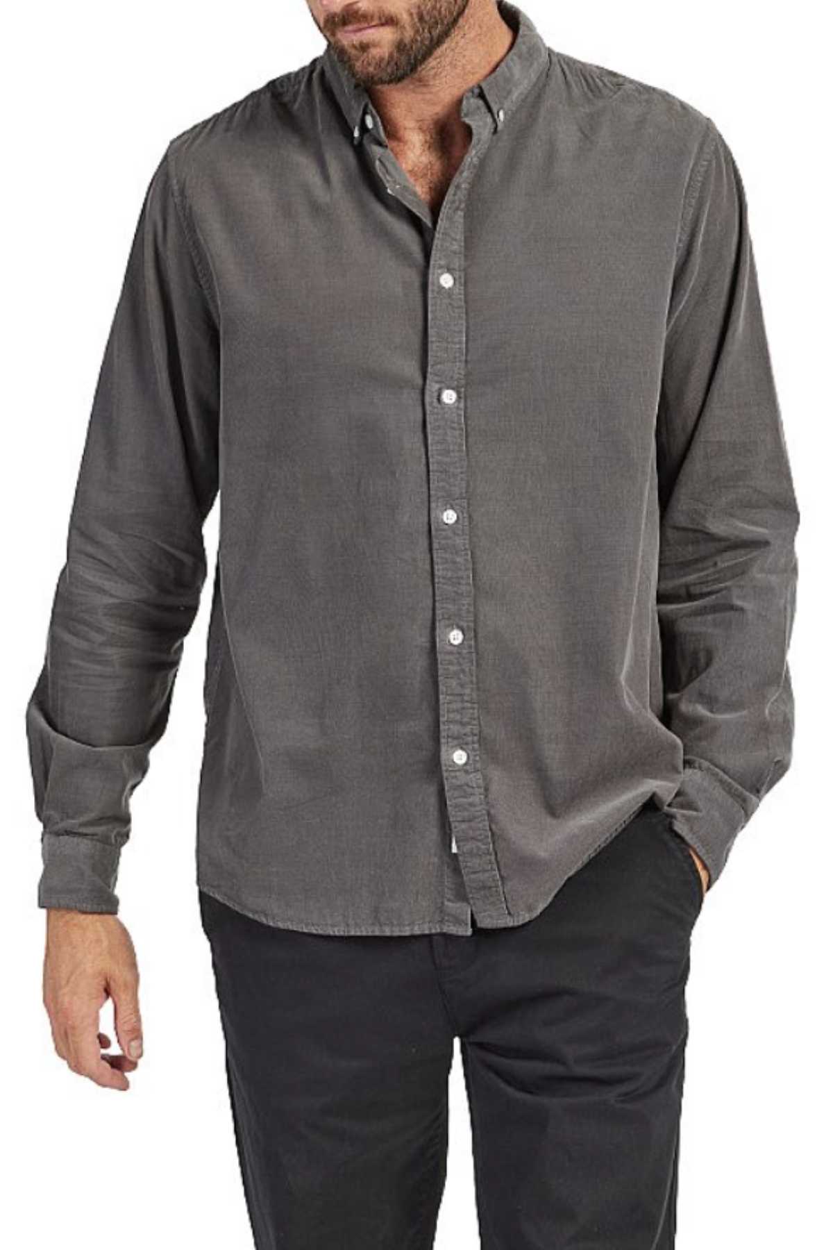 A man is wearing a grey, long-sleeved Academy Brand Micro Cord Shirt made from 100% cotton and black pants. His left hand is in his pocket, and his right arm hangs by his side. He has a slight beard and short hair, though his face is partially out of frame, giving him a polished look.