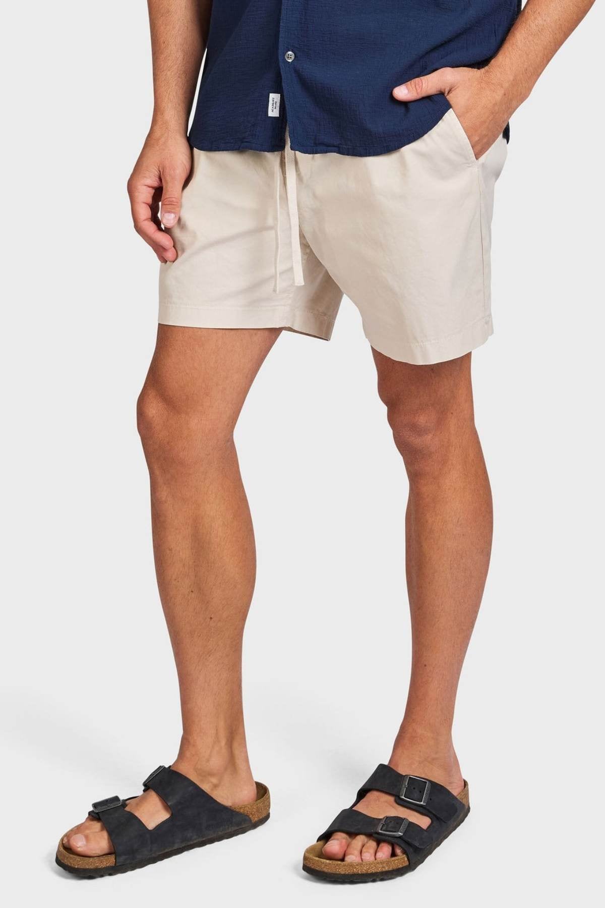 A person wears a navy blue shirt, Academy Brand Oahu Short cotton twill shorts, and black sandals with cork soles, standing with one hand in their pocket against a plain background.